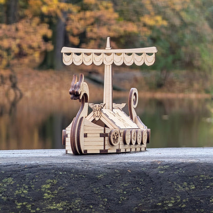 Laser cut wood Viking longship model resting on a weathered wooden board outdoors, showing curved prow and patterned shields.