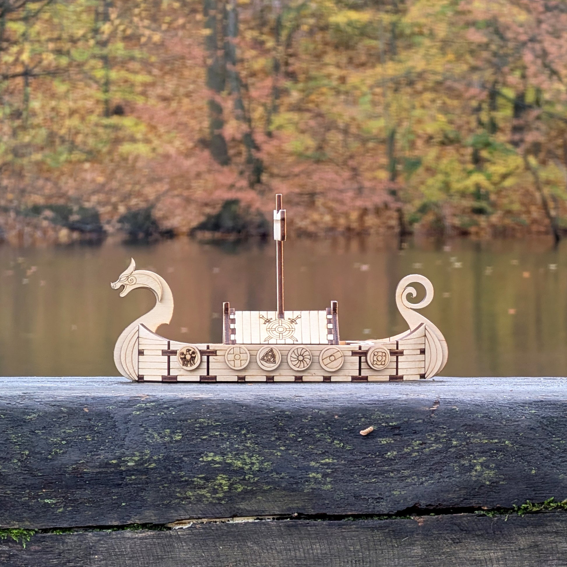 Side view of a laser cut wooden Viking longship model photographed in front of a lake with colorful fall foliage behind it.