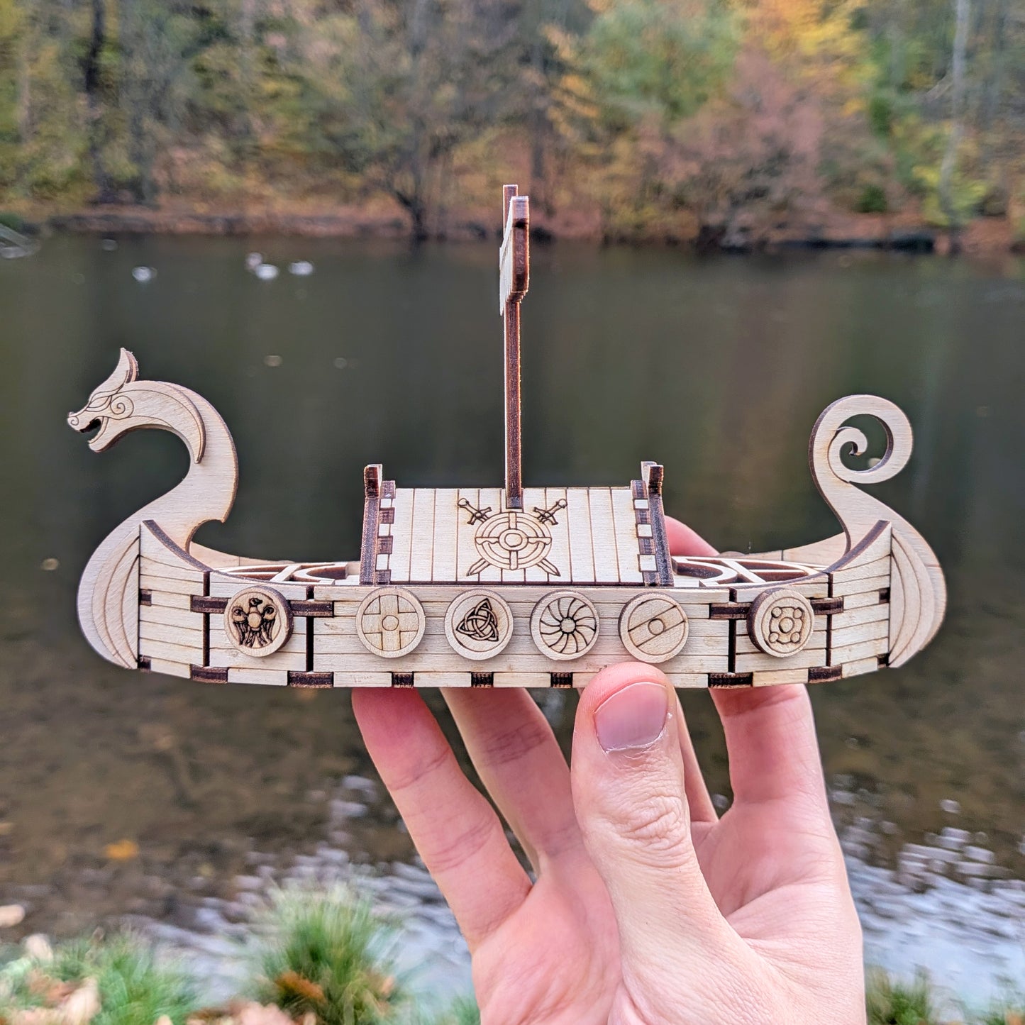 Side profile of the laser cut wooden Viking ship model with dragon head and curled stern, photographed near water in autumn.