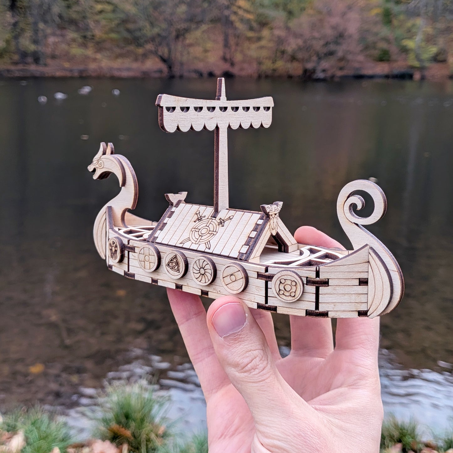Laser cut wooden Viking ship model held in a person's hand to show scale, with a blurred lake background.