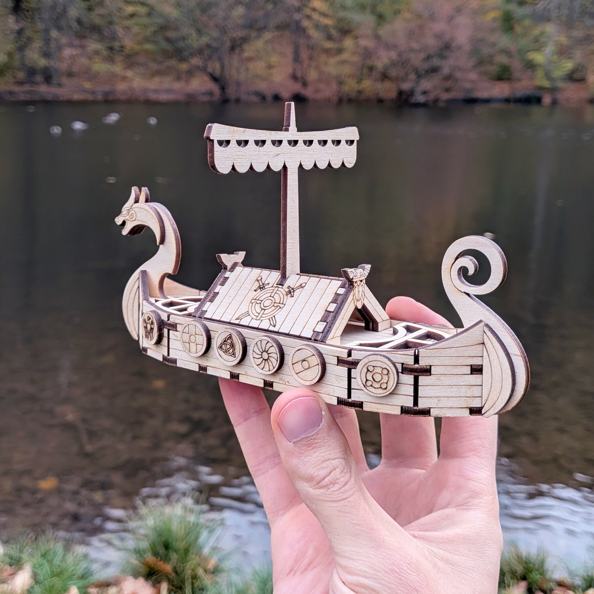 Laser cut wooden Viking ship model held in a person's hand to show scale, with a blurred lake background.