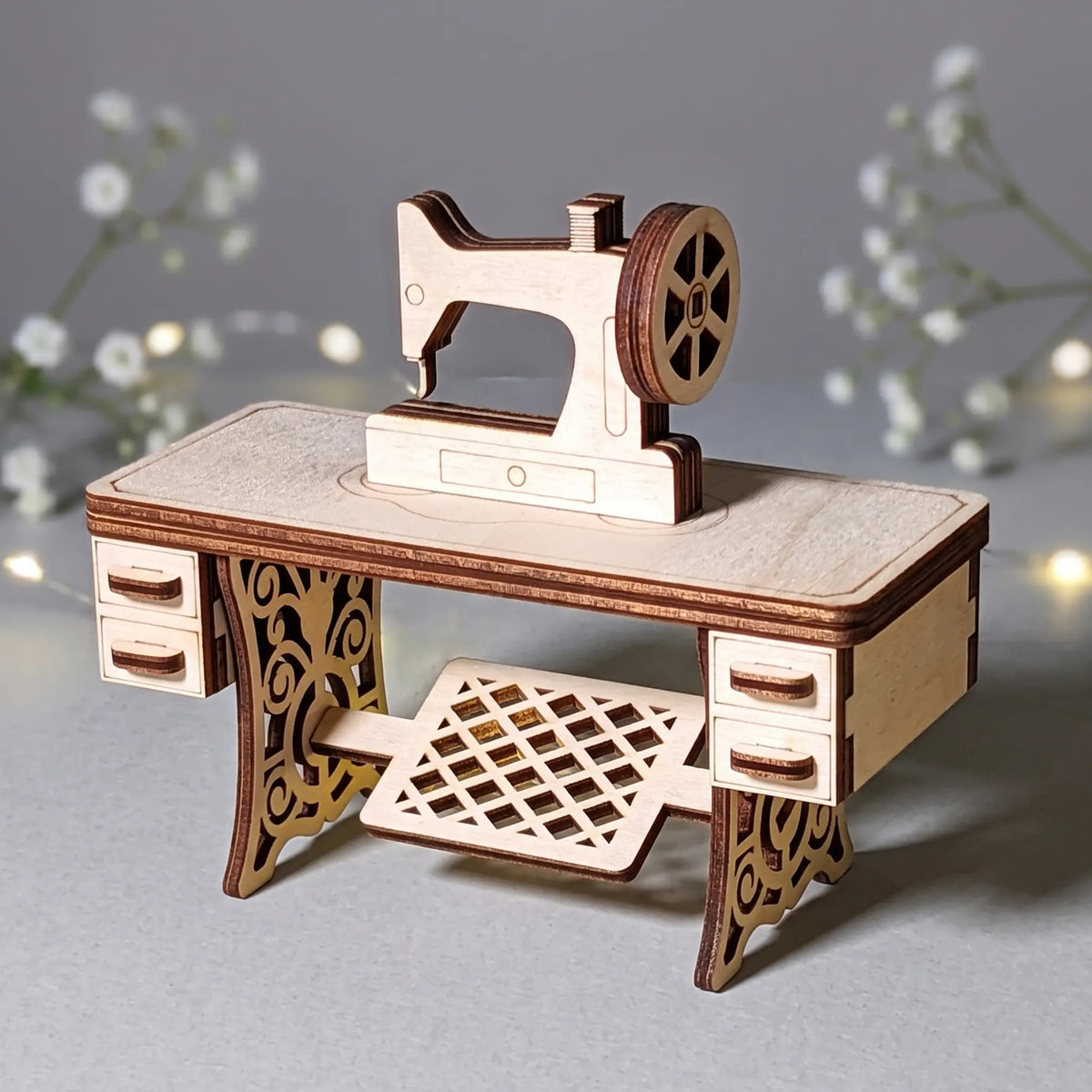 A wooden sewing machine laser cut model is displayed on a desk, with a background of white flowers and twinkling lights.