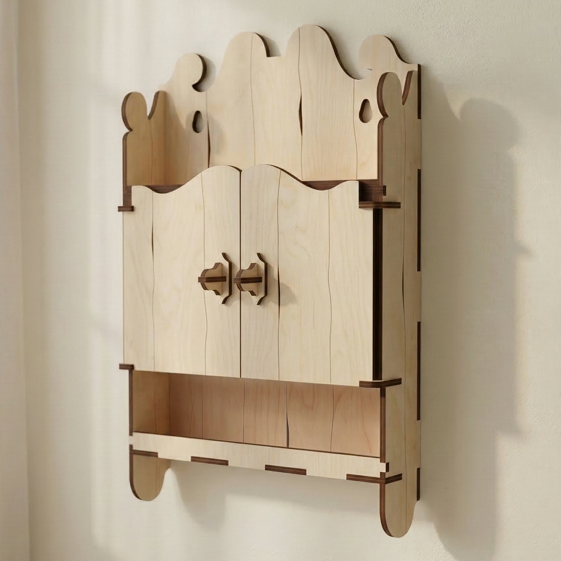 Miniature ornate cabinet made of laser cut plywood with two closed doors and an open bottom shelf. Features a decorative scalloped top. Natural light wood texture with dark edges, displayed on a white table next to a coffee mug.