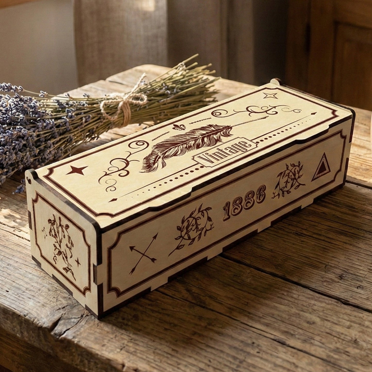 Rectangular laser cut plywood box with a vintage aesthetic. The lid is engraved with a large feather and filigree. Side panels show "1886" and floral motifs. Placed on a rustic table with dried lavender and a ceramic mug for scale.