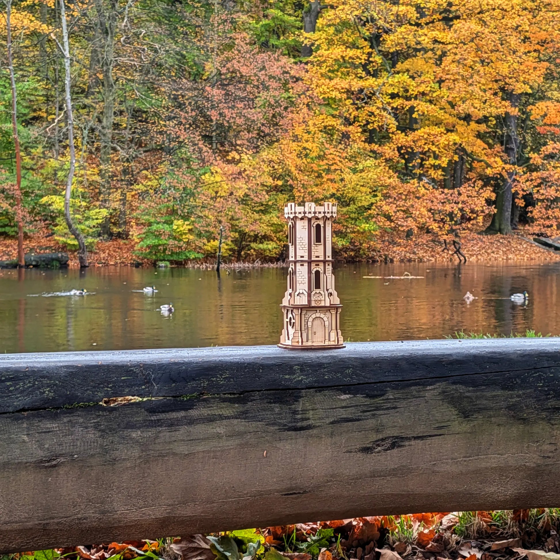 A distant scenic view of a miniature medieval watchtower model in laser cut plywood on a wooden fence near an autumn lake