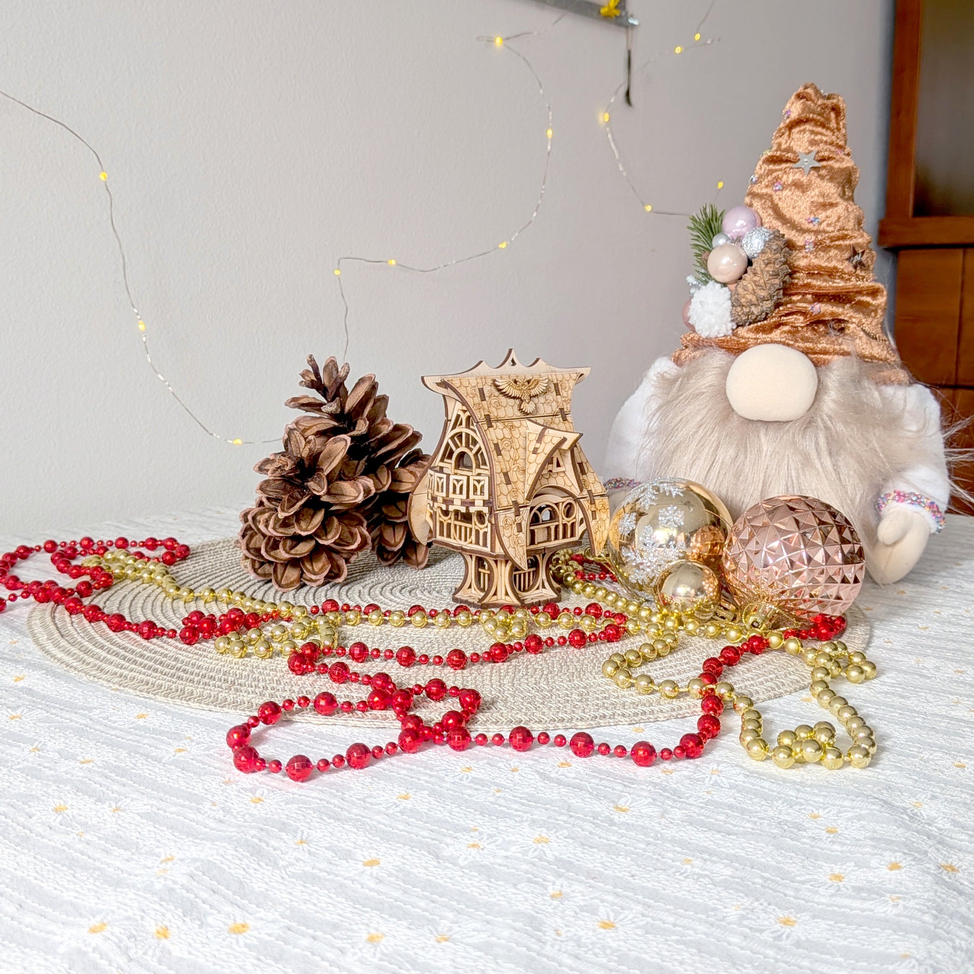 Laser cut wooden fairy house styled in a holiday scene with pinecones, ornaments, and red and gold beads on a light woven surface.