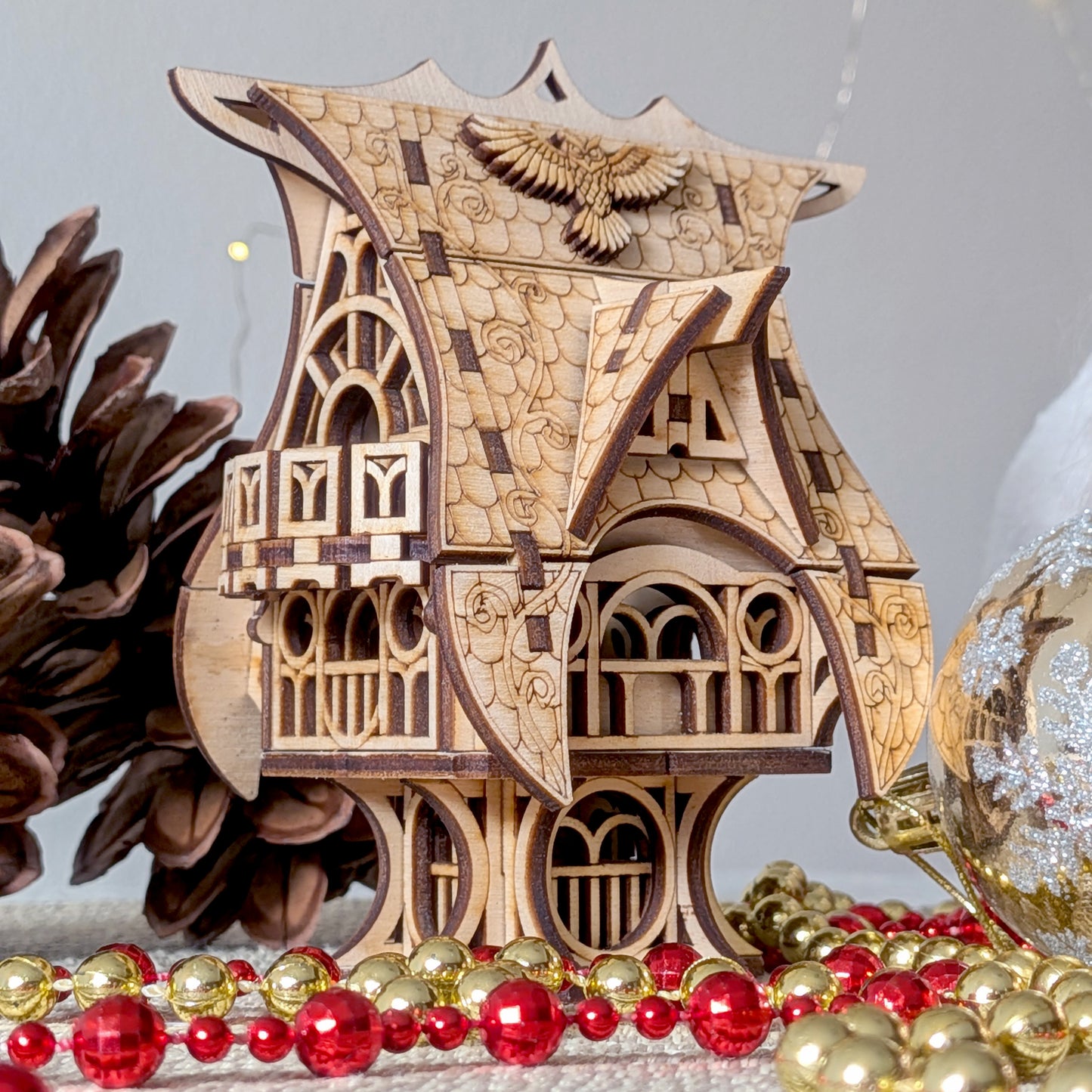 Close front view of the laser cut wooden fairy house with pinecones and holiday ornaments, showing the owl roof detail and curved wooden structure.