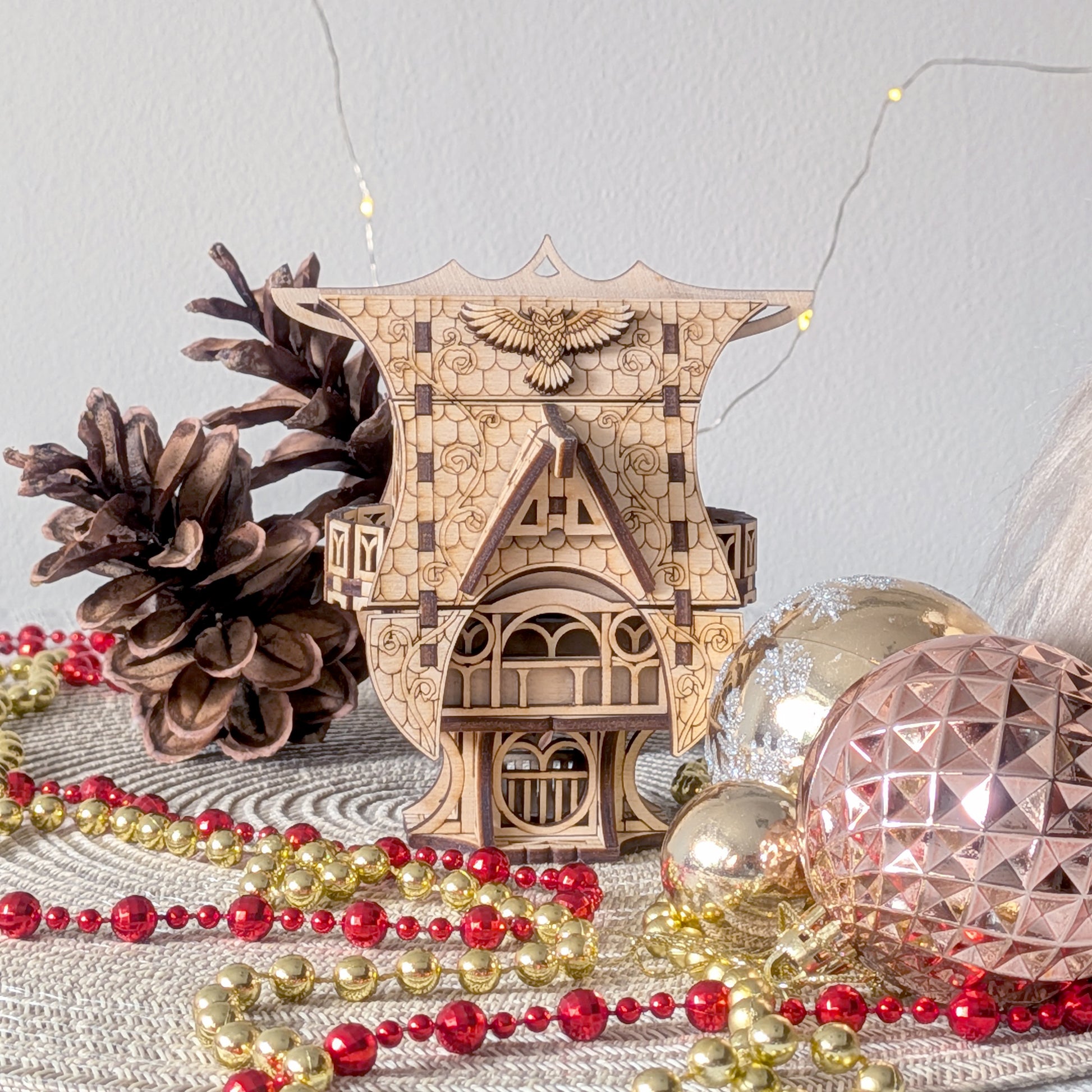 Front view of the laser cut fairy house decorated for the holidays, with pinecones and shiny ornaments arranged around it.