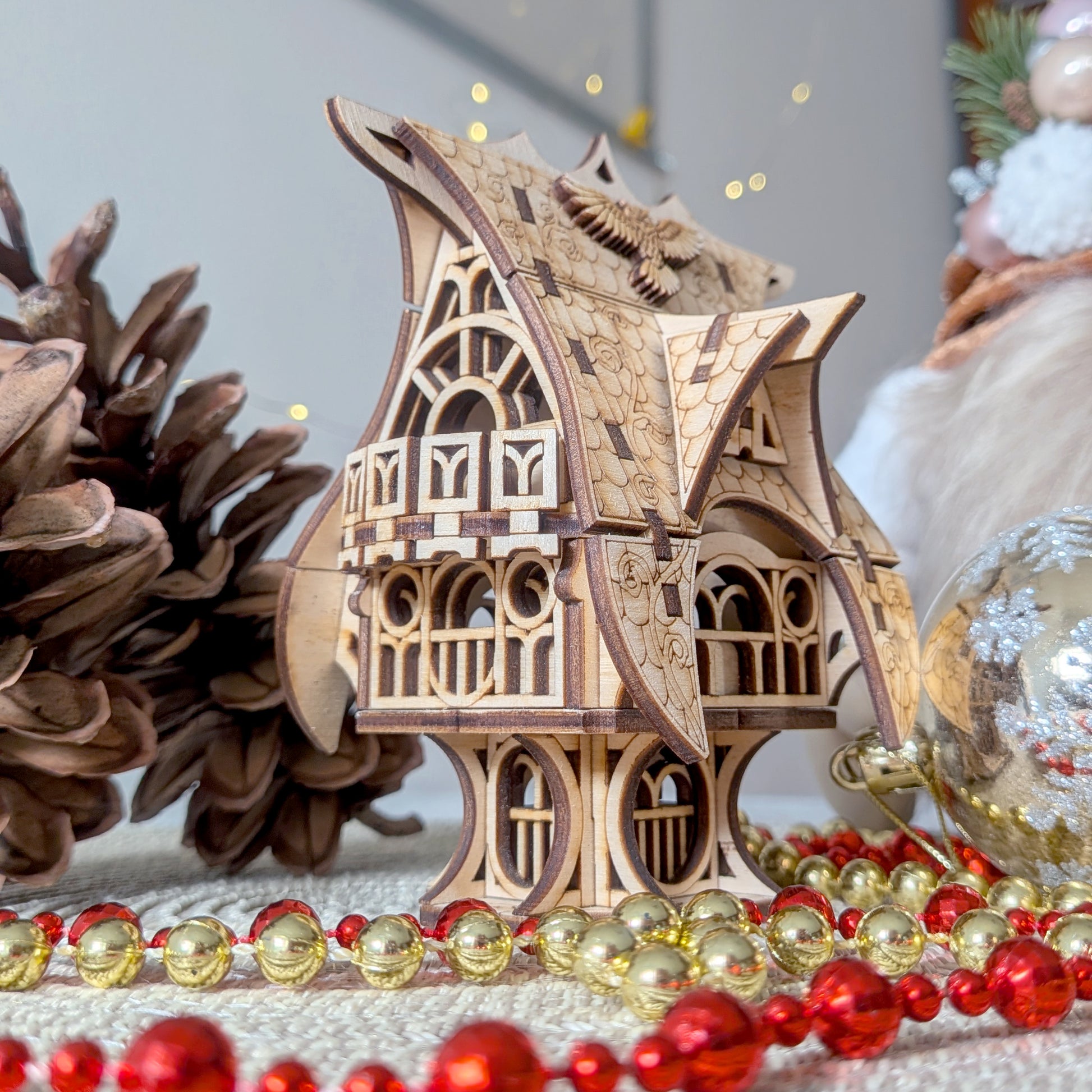 Laser cut wooden fairy house model displayed with pinecones and holiday ornaments, showing detailed engraved shingles and curved roof accents.