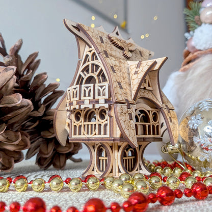 Laser cut wooden fairy house model displayed with pinecones and holiday ornaments, showing detailed engraved shingles and curved roof accents.