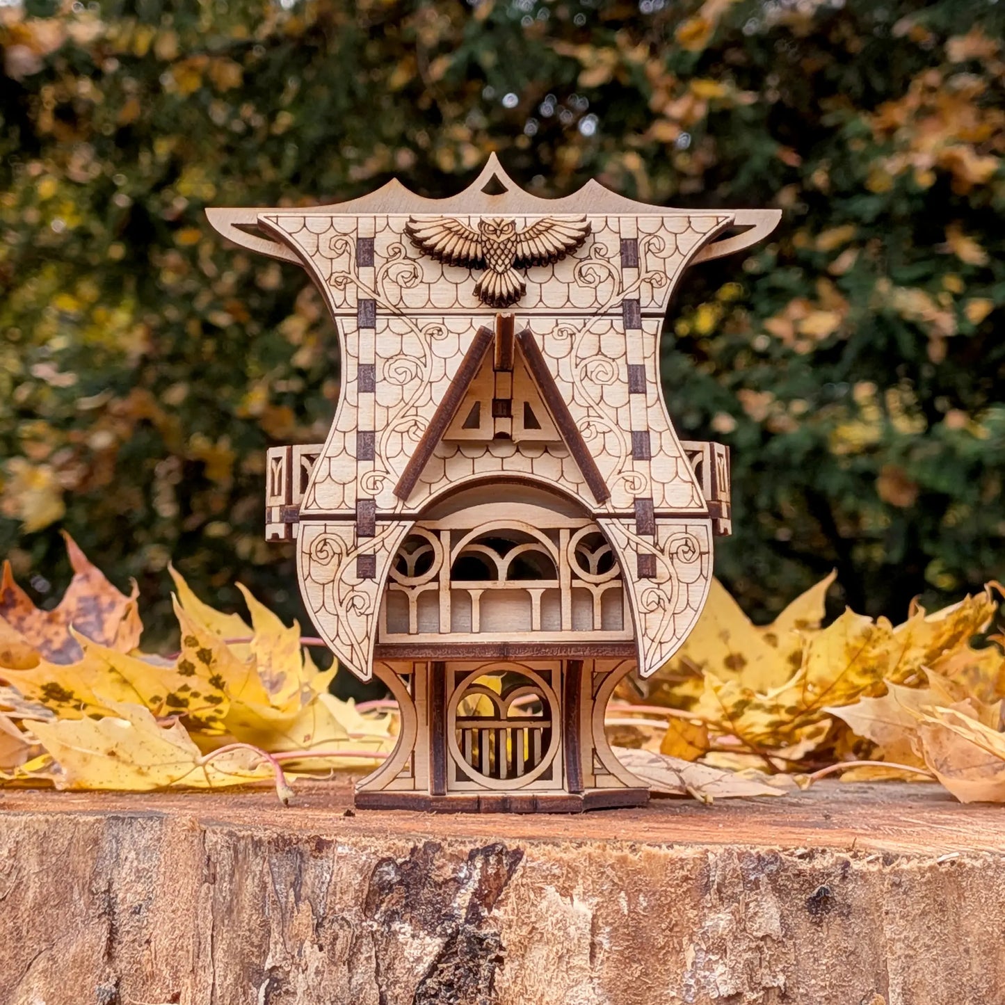 A clean straight front view of a miniature whimsical fairytale owl cottage of laser cut plywood upon a forest tree stump