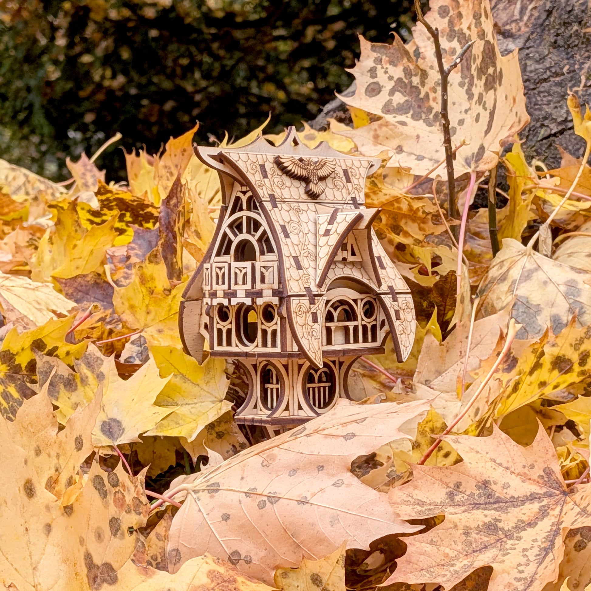 Laser cut wooden fairy house nestled among fallen autumn leaves, emphasizing its detailed engraving and whimsical cottage shape.