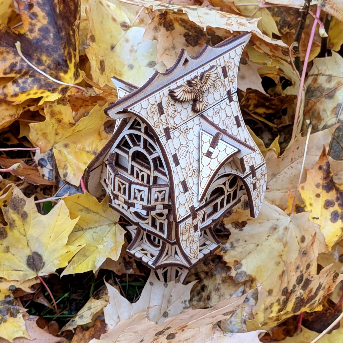 A sharp high-angle view looking down at a miniature fairytale owl cottage in laser cut plywood inside dense autumn leaves