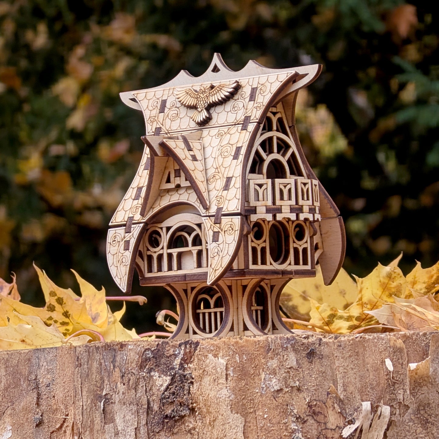 Laser cut wooden fairy house shown outdoors on a tree stump surrounded by autumn leaves, highlighting its carved windows and curved roof.
