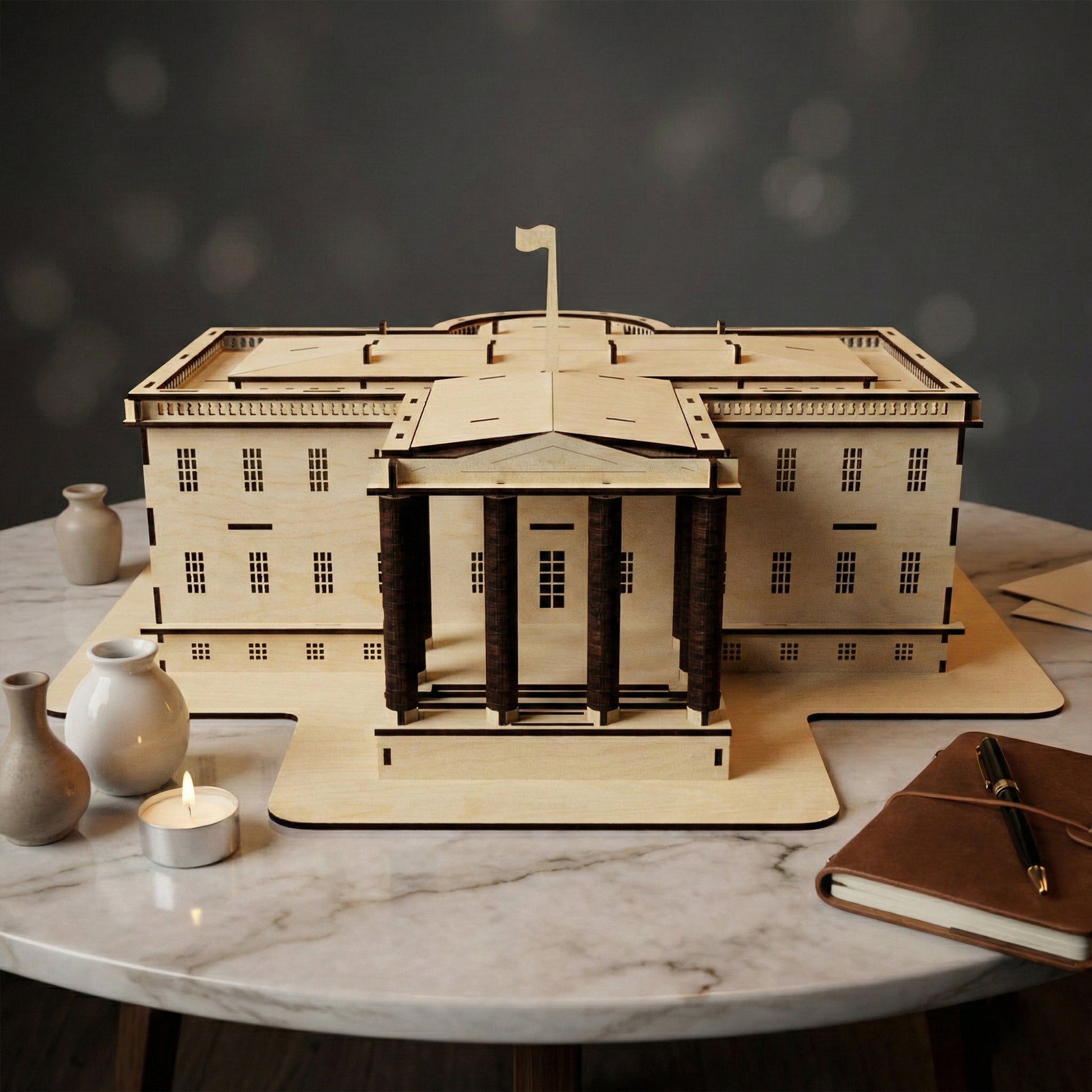 Frontal view of a White House model made from laser cut plywood. Features a prominent gabled portico with dark wooden columns. Rests on a marble table next to a burning candle, small ceramic vases, and a leather notebook.