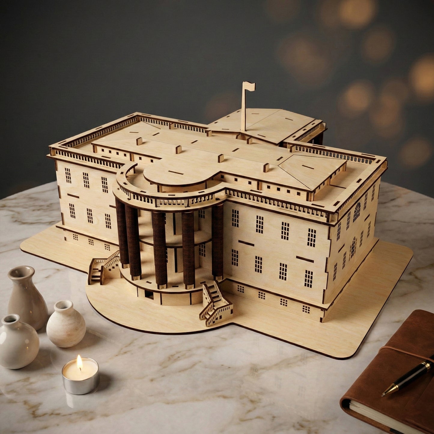 Perspective view of the laser cut plywood White House, highlighting the 3D construction and layered roof details. The natural wood grain is visible. Placed on marble with a leather journal and pen in the foreground for scale.