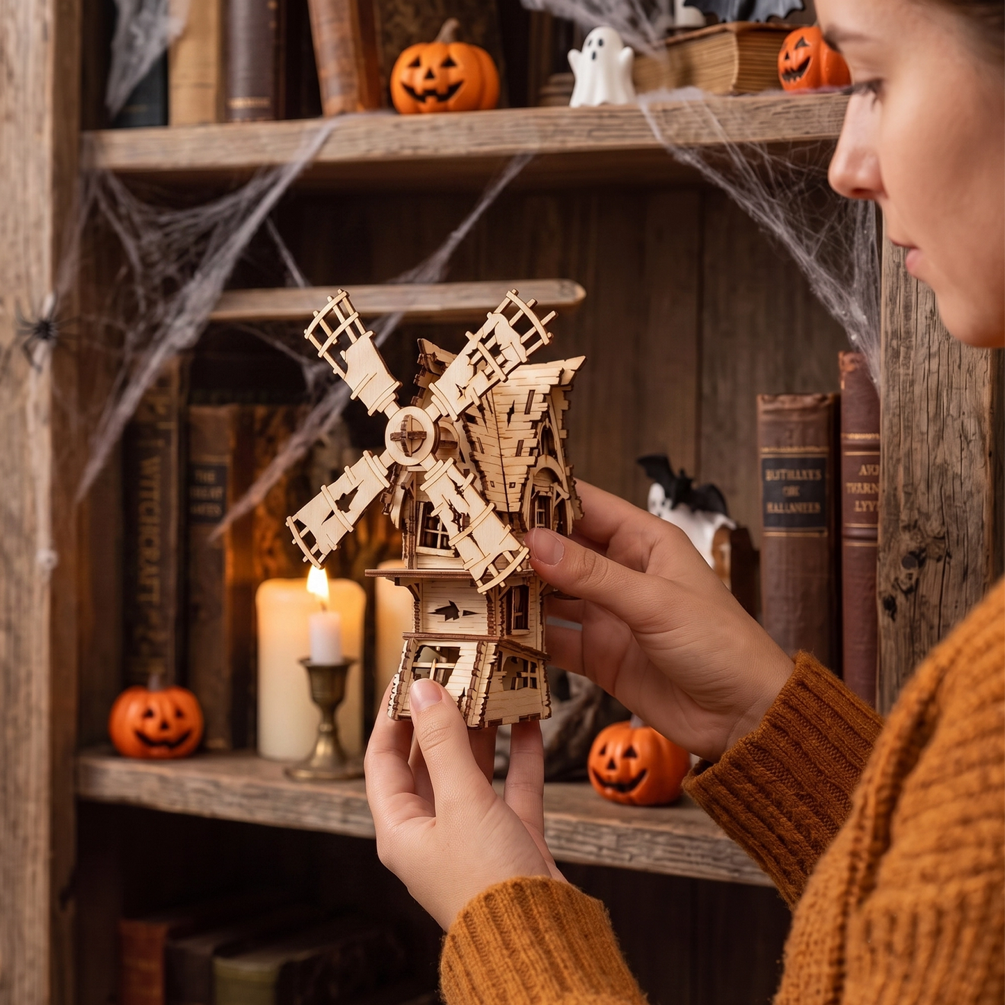 Hands holding a laser-cut birch plywood Halloween windmill model in a spooky bookshelf setup with pumpkins and candles