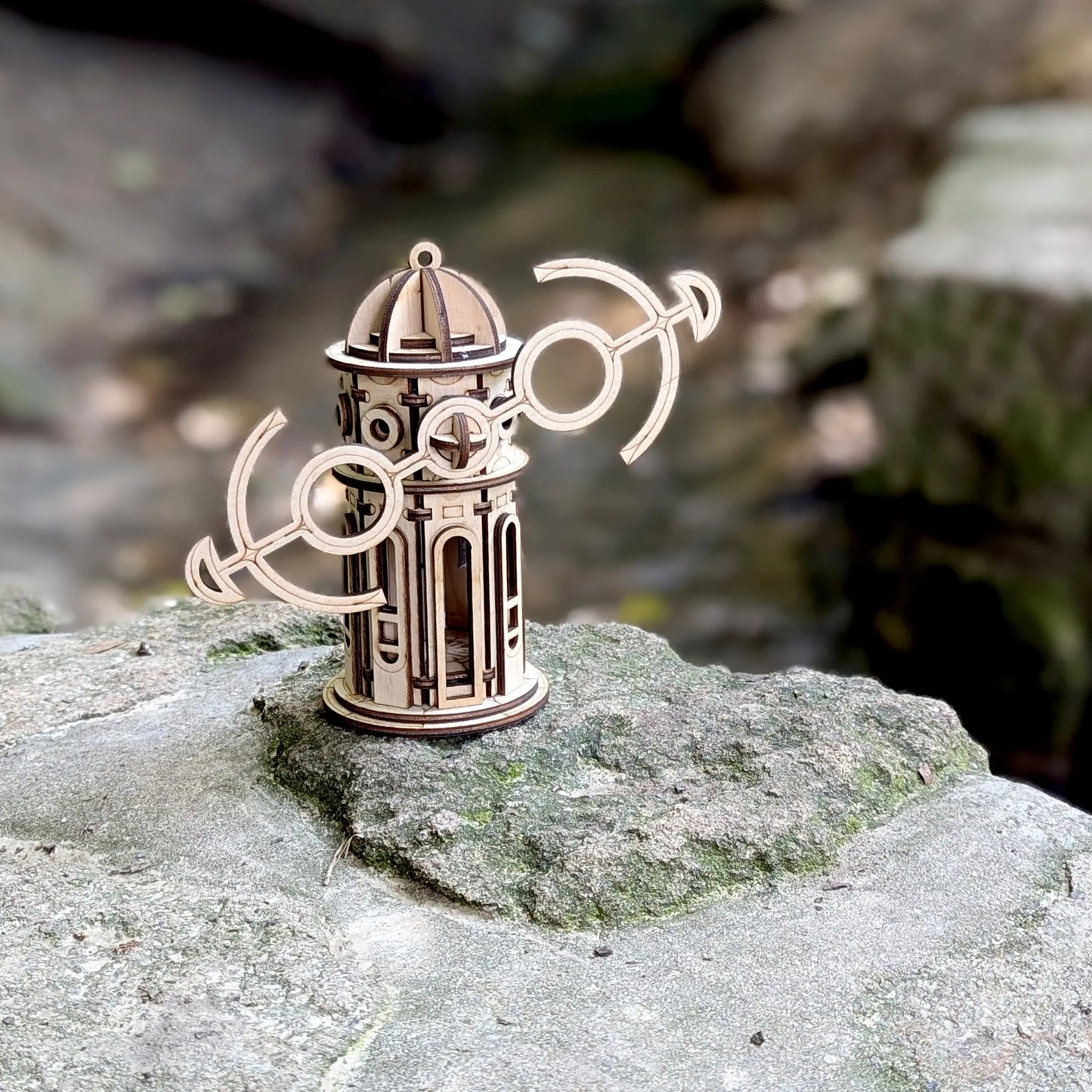 Laser cut plywood kinetic windmill toy with anchor wings tilted right, standing on a grey mossy rock near a creek.