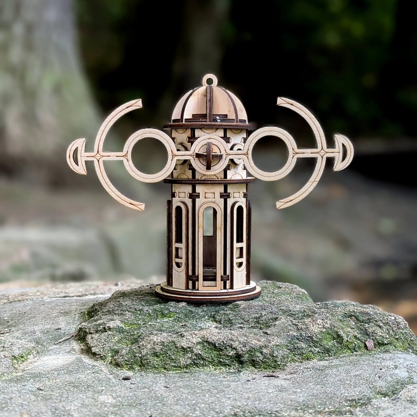 Laser cut plywood kinetic windmill ornament with horizontal anchor wings, standing on a large mossy rock.