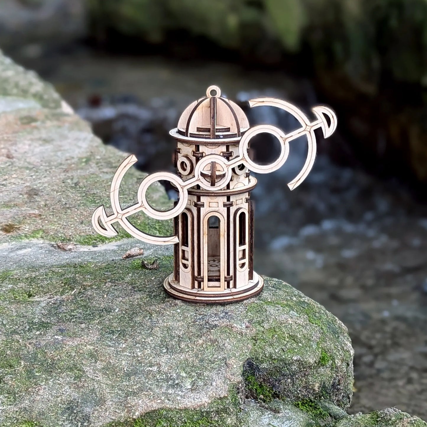 Miniature laser cut plywood windmill ornament with tilted anchor wings, standing on a mossy stone by a river.