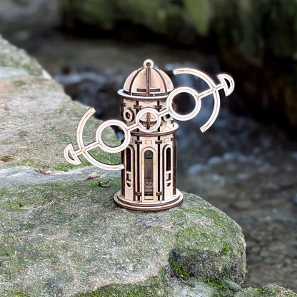 Miniature laser cut plywood windmill ornament with tilted anchor wings, standing on a mossy stone by a river.
