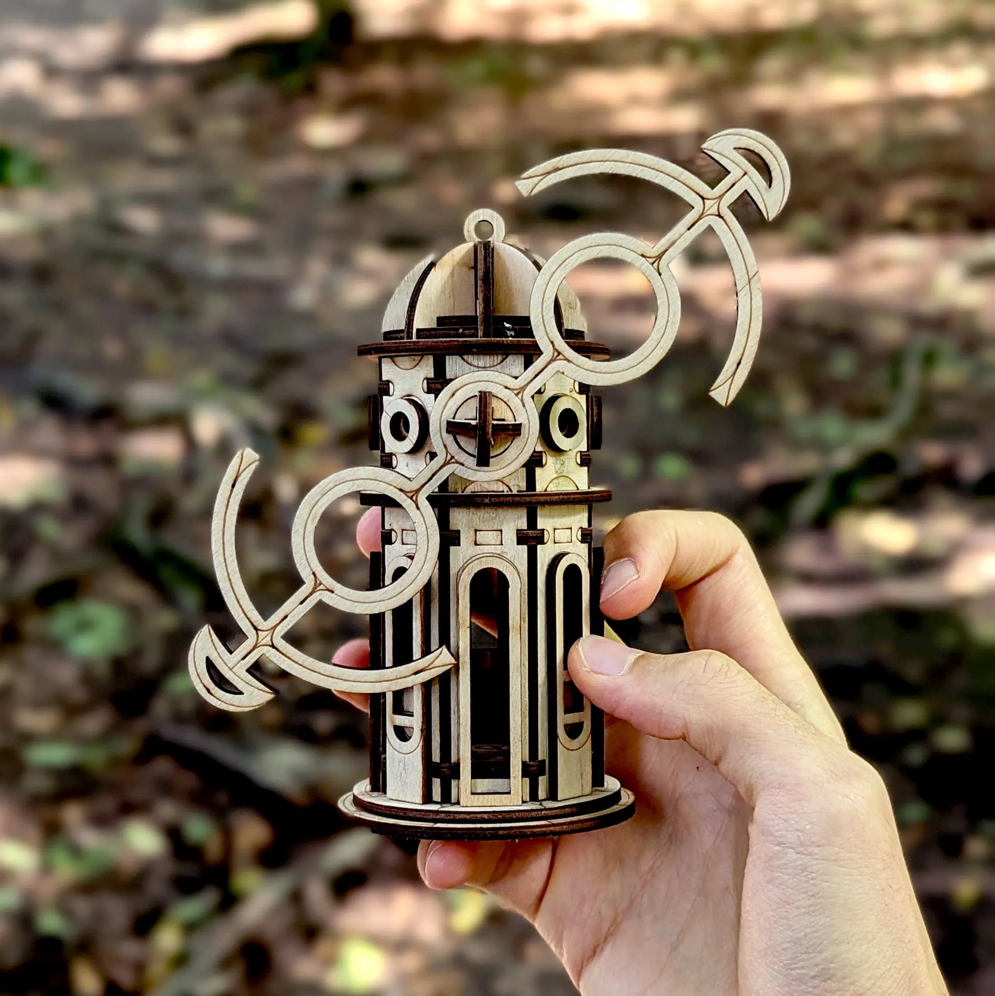 Human hand holding a small laser cut plywood windmill tower with tilted anchor shaped wings in a blurry forest.