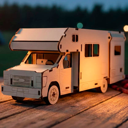 Side view of a laser cut wooden plywood truck camper model with a slide-out section, resting on a picnic table. The 3D plywood puzzle features intricate window cutouts and wheel details, set against a twilight campsite with a red plaid blanket