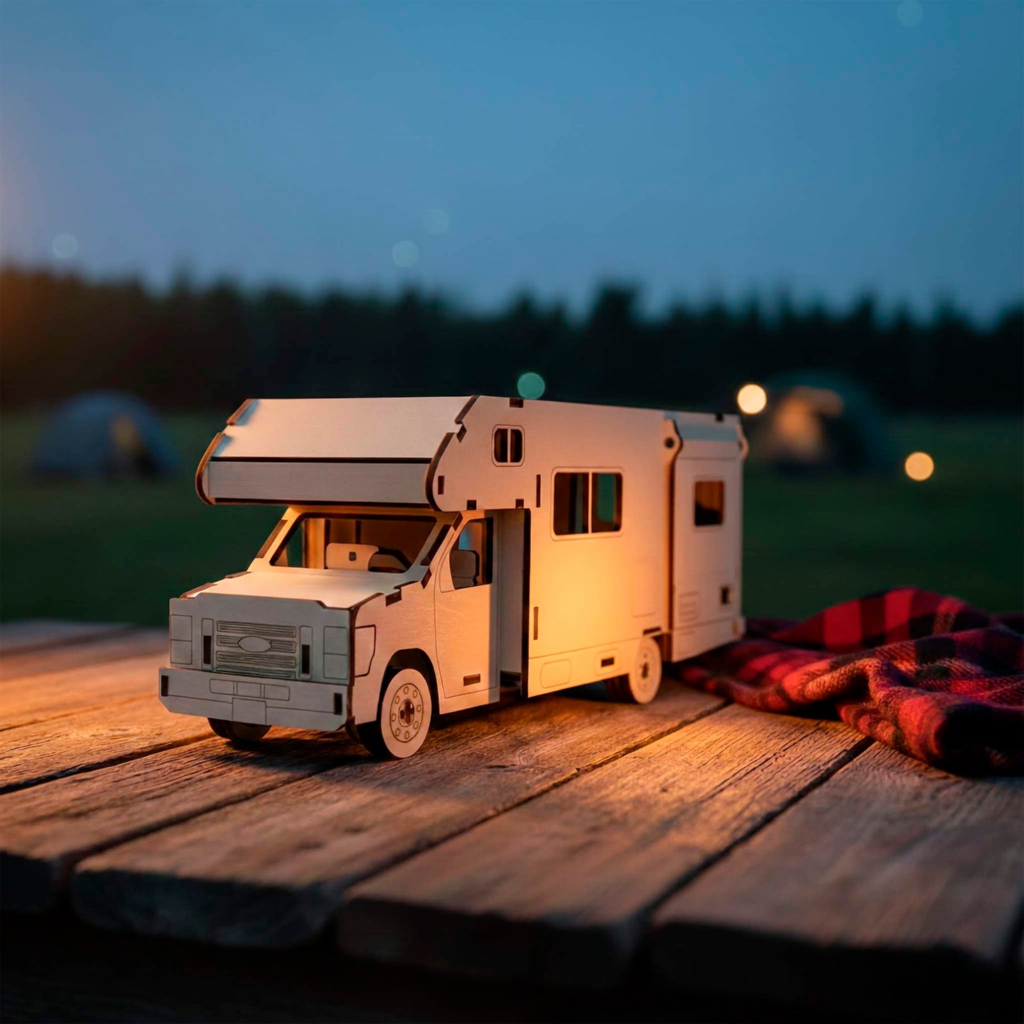 Side view of a laser cut wooden plywood truck camper model with a slide-out section, resting on a picnic table. The 3D plywood puzzle features intricate window cutouts and wheel details, set against a twilight campsite with a red plaid blanket