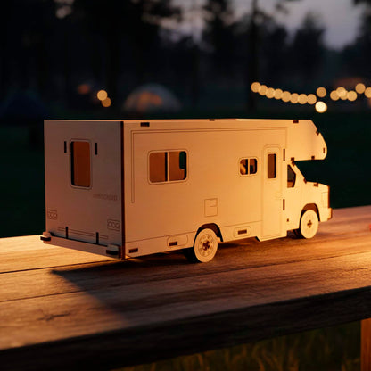 Rear view of a laser cut wooden plywood Winnebago camper model on a wooden table at night. Detailed plywood construction showing the back door, windows, and "OUTLOOK" engraving, illuminated by warm evening light with blurred tents in the background.