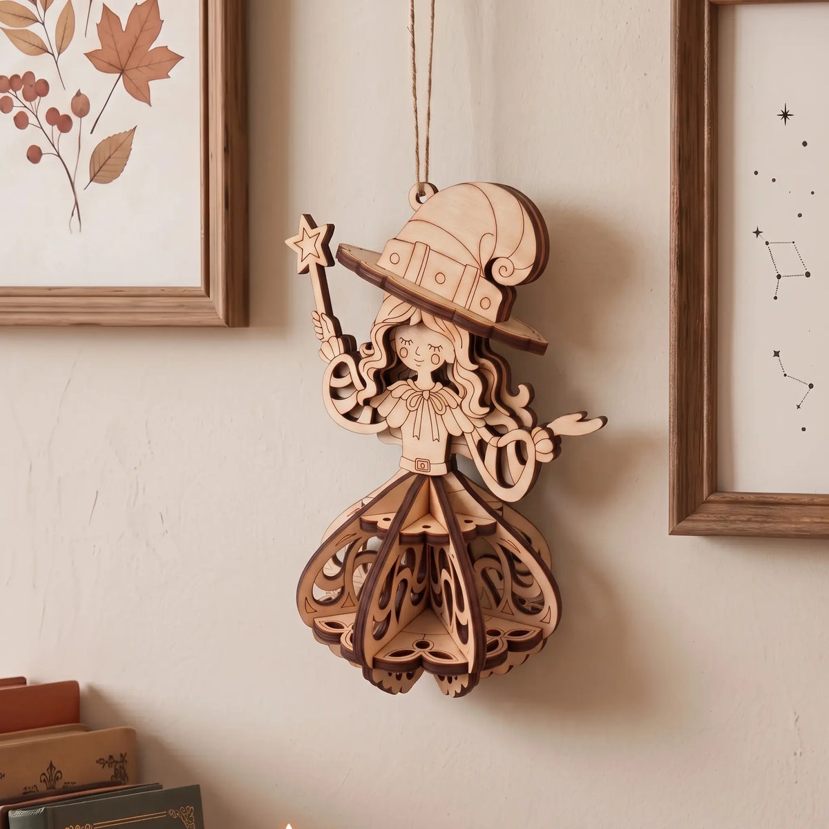 A laser-cut wooden hanging ornament shaped like a whimsical fairy or witch with a tall hat, wand, and layered skirt, displayed on a beige wall between framed botanical and starry prints.