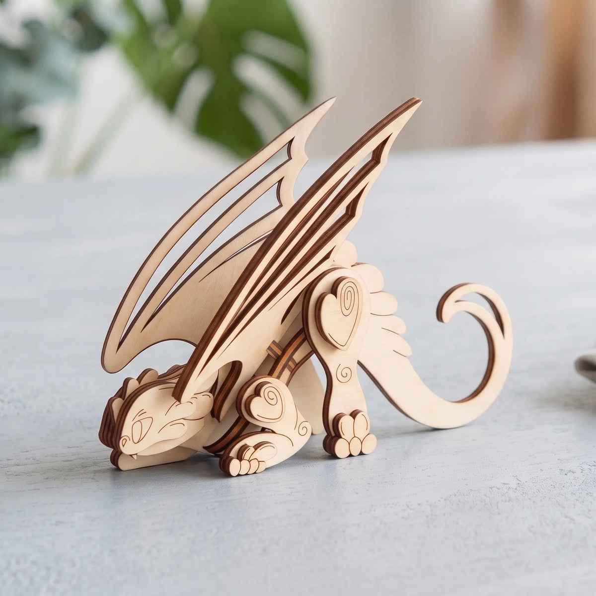 3D laser cut plywood crouching dragon figurine with spread wings on a light grey tabletop near blurred indoor houseplant.