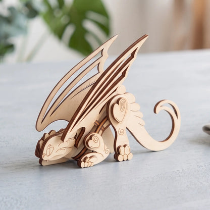 3D laser cut plywood crouching dragon figurine with spread wings on a light grey tabletop near blurred indoor houseplant.